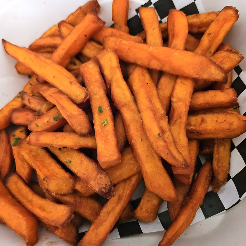 A close-up of crispy sweet potato fries, some with green herb flecks, in a black and white checkered paper container.